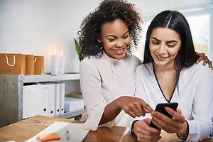 Two women are smiling and looking at a cell phone in an office room.