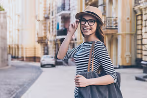 A smiling woman wearing glasses and a hat stands on a city street, holding a bag and looking away.