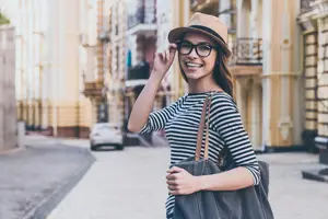 A smiling woman wearing glasses and a hat stands on a city street, holding a bag and looking away.