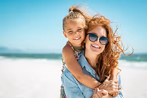 A woman and a girl are smiling on the beach with the ocean in the background.
