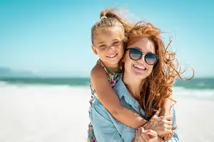A woman and a girl are smiling on the beach with the ocean in the background.