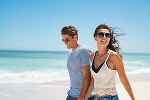 A man and a woman walking on the beach under a blue sky with small waves in the distance