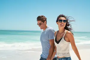 A man and a woman walking on the beach under a blue sky with small waves in the distance