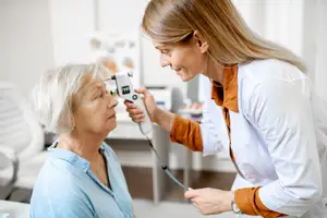 An optometrist in a white lab coat is checking the eyes of a senior woman with a slit lamp