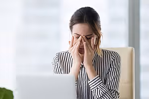 A woman sitting in an office chair covering her face with her hands while looking at her laptop