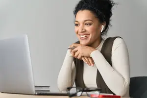 A smiling woman wearing headphones and sitting at a desk with a laptop, books, and eyeglasses