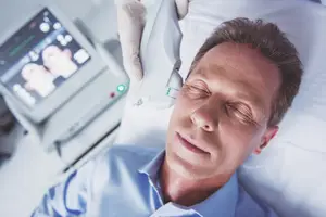 A man is lying on a bed in a hospital room, possibly undergoing an ultrasound procedure, with a monitor in front of him displaying a picture of a baby and the man wearing a blue polo shirt with a white glove on his hand, holding a device.