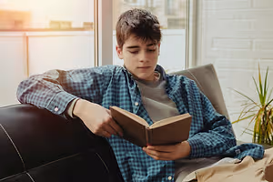 A boy with short brown hair is sitting on a couch reading a book, his eyes closed with his head leaning against the wall. He is wearing a blue checkered long-sleeve shirt and a gray shirt underneath. He is holding a brown book with his right hand, while his left hand is resting on the book. A plant is on the right side of the couch, and a glass window is behind the boy. Behind the window, there are buildings with brick walls.
