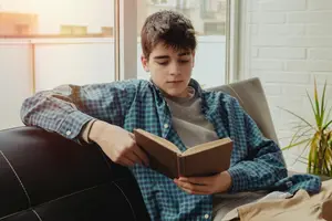 A boy with short brown hair is sitting on a couch reading a book, his eyes closed with his head leaning against the wall. He is wearing a blue checkered long-sleeve shirt and a gray shirt underneath. He is holding a brown book with his right hand, while his left hand is resting on the book. A plant is on the right side of the couch, and a glass window is behind the boy. Behind the window, there are buildings with brick walls.