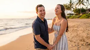 A man and woman stand on the beach at sunset, both smiling and holding hands.