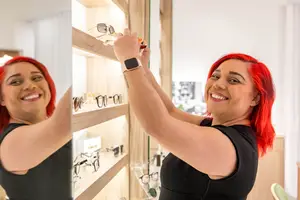 A woman with red hair is smiling and fixing a pair of glasses on a wooden shelf in a shop. She is wearing a black sleeveless top and a watch.