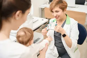 A doctor is examining a baby in a clinic with a smiling mother and a medical professional holding an eye examination tool.