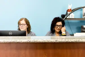 Two women are sitting at a reception desk, one is using a laptop and the other is on a phone call.
