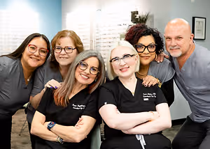 A group of smiling people, including two women wearing glasses, are posing for a photo in a room with a blue wall and a picture frame.