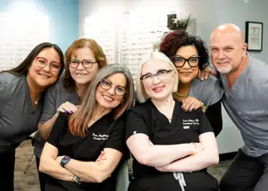 A group of smiling people, including two women wearing glasses, are posing for a photo in a room with a blue wall and a picture frame.