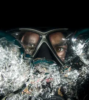 A person wearing a scuba mask is underwater, with water droplets around them and their reflection visible in the mask.
