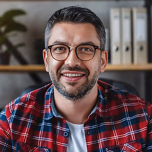 A smiling man wearing glasses and a red plaid shirt sitting in an office chair.