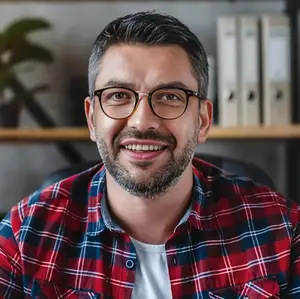 A smiling man wearing glasses and a red plaid shirt sitting in an office chair.