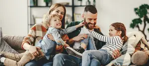 A family of four sitting together on a couch in a cozy living room setting, smiling and laughing, with the father holding the child and the mother holding the other child.