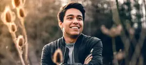 A smiling man in a black jacket stands with folded arms in a forest with a bright light illuminating his face