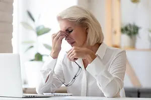 A woman sitting at a desk with a laptop and potted plants, she is holding her face with one hand and her glasses with the other, looking tired or stressed.