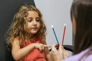 A young girl with curly hair is pointing at something while a woman in a purple shirt holds two red and blue rattles, possibly in a medical setting.