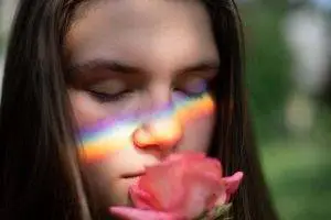 A woman with a rainbow painted on her face and eyes closed smelling a rose
