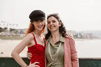 Two women standing together smiling for a photo with a view of the beach and buildings behind them.