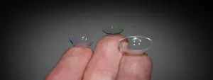 Two transparent colored contact lenses are held by a fingertip against a gray background.