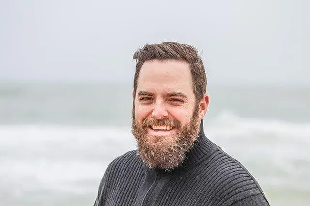 A man with a beard smiling on the beach with waves in the background