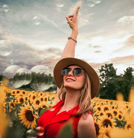 A woman standing in a sunflower field wearing a hat and sunglasses and pointing up at bubbles in the sky.