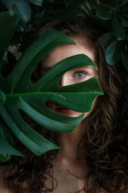 A woman with curly hair hides behind a large green leaf, showing her eyes and lips