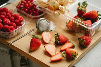 Various fresh fruits are arranged on a wooden chopping board, including strawberries, raspberries, and what appears to be a flower. A strainer is also placed on the board, suggesting the preparation of a fruit-based dish.
