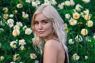 A blonde woman with long hair stands in a field of yellow and white flowers, smiling for a photo.