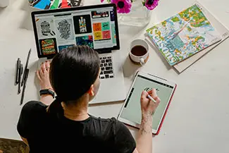 A person sitting at a desk using a laptop and a tablet, wearing a black shirt, with various office items on the desk, like books, pens, and a cup.