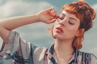 A woman with red hair is wearing a pink and white printed shirt and is blowing her nose with her hand while looking down with her eyes closed.