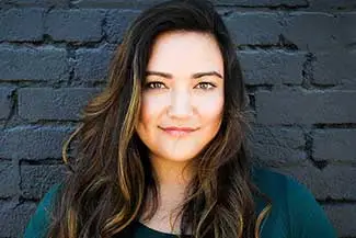A smiling woman with long curly hair stands in front of a brick wall.