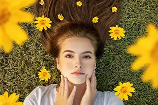 A woman with long hair is lying on the grass with yellow flowers around her.
