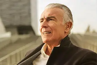 A smiling older man with white hair and a mustache, wearing a black jacket and white shirt, sitting on a bench in an outdoor area, looking to the right with a smile on his face.