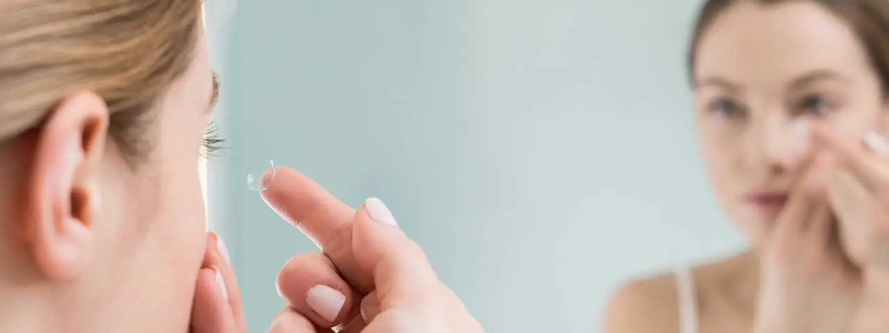 Woman placing a contact lens into her eye in front of a mirror