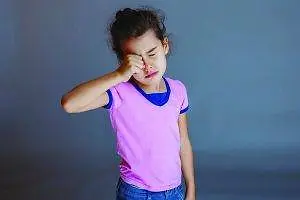A girl in a pink shirt and blue jeans is holding her nose with her right hand and seems to be crying in a room with a gray wall.