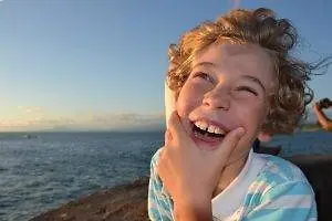 A young boy with curly hair is smiling while standing near a body of water, possibly the sea, and posing for a photo. He has his hand on his cheek and seems to be looking at something in front of him. The sea has calm water and is reflecting the sky with clouds. In the distance, there is a silhouette of a person taking a photo. 