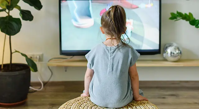 A little girl is watching television while sitting on a stool in a room with a TV, potted plants, and a shelf with a disco ball.
