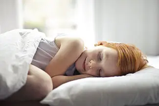 A redheaded girl sleeps peacefully in her bed with sunlight streaming in through a window