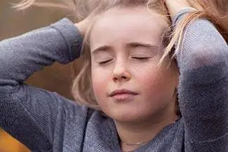 A girl with her eyes closed, holding her head with both hands. She is in a forest, probably resting after a long day.