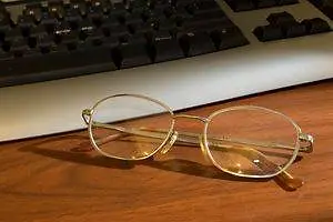 A pair of gold-rimmed glasses with a shadow cast on a wooden desk, next to a black keyboard