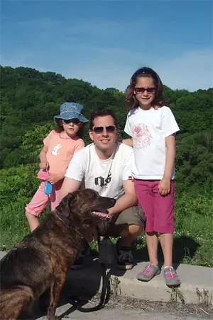 A man and two girls are smiling and posing for a photo with a dog and a landscape in the background.