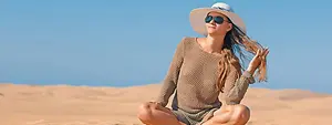 A woman sitting on the sand in the desert with sunglasses and a hat