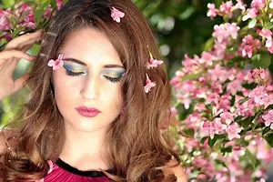 Woman with brown hair and floral makeup poses in front of pink flowers