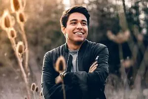 A smiling young man stands in a field of tall, dried grass with a blurred background of trees and bright sunlight shining through the trees